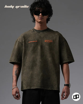 Person wearing a 'Behind For A Reason' Olive Green 240 GSM 100% Cotton Oversized Acid Wash T-Shirt with brand logos on a gray background - Front