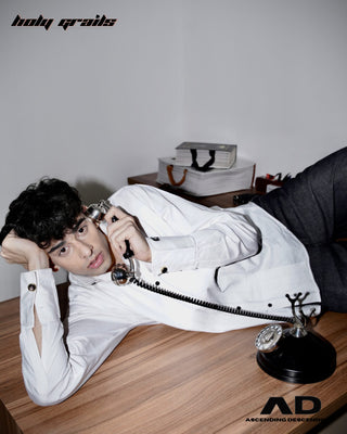 Men lying on a wooden table with a vintage phone, wearing a street style 'Cuff Script' White Pintex Cotton Pleated Designer Shirt - Side
